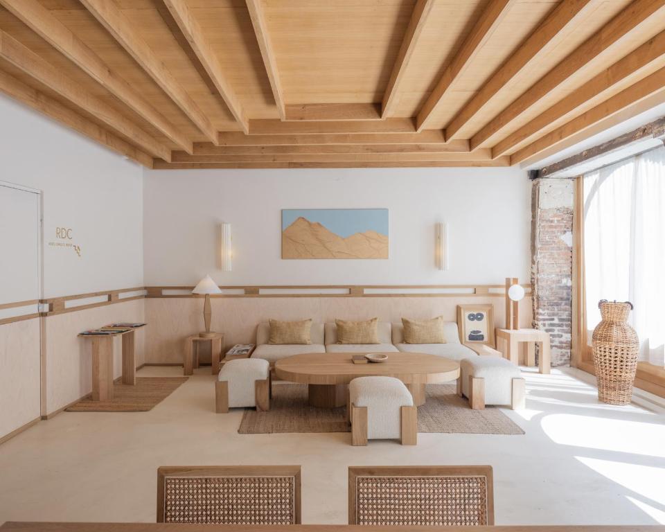 13 Unique Hotels in Paris for a Stylish Stay 25 A modern living room reminiscent of unique hotels in Paris, with light wood beams, neutral furniture, low round tables, beige cushions, a woven rug, abstract art, large windows with sheer curtains, and an exposed brick wall flooded by natural light.