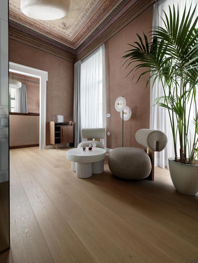A stylish living room with tall windows, sheer white curtains, light wood flooring, and modern round chairs evokes the chic ambiance found in the best boutique hotels in Athens. A potted plant and intricate ceiling molding add refined charm.
