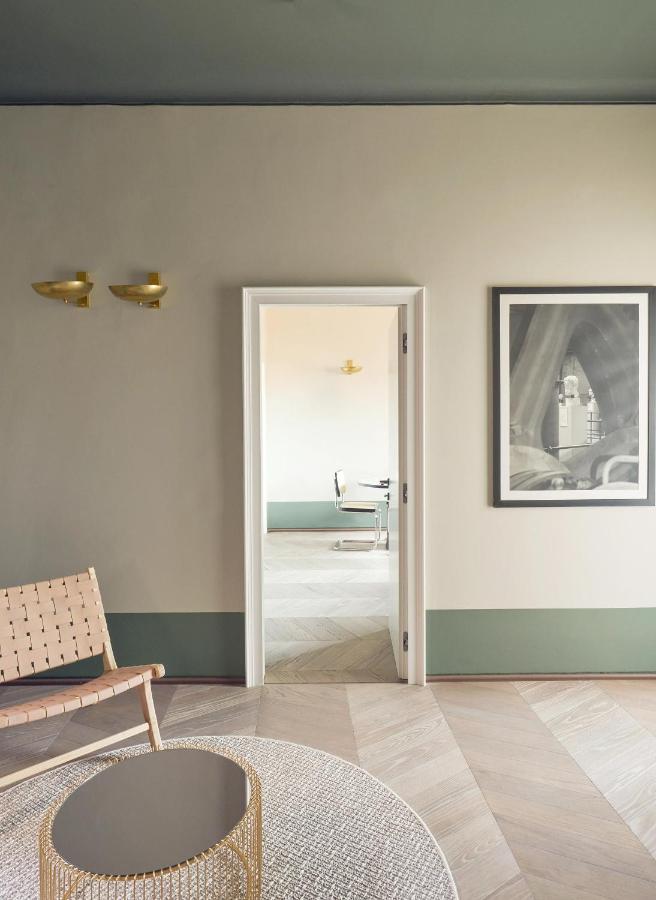 13 UNIQUE Boutique Hotels in Rome 18 A minimalist, modern interior reminiscent of boutique hotels in Rome, Italy, features muted green and beige walls, a wooden chair, round coffee table, framed artwork, and an open door revealing a bright room with a table and chair.