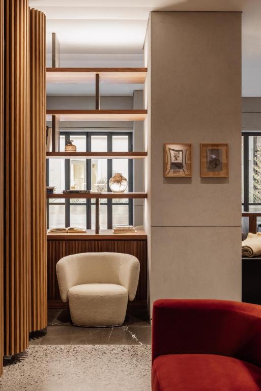 A cozy modern interior, reminiscent of boutique hotels in Athens, with a cream armchair before wooden shelves, a partial view of a red sofa, framed art on the wall, large windows, and decorative objects on the shelves.
