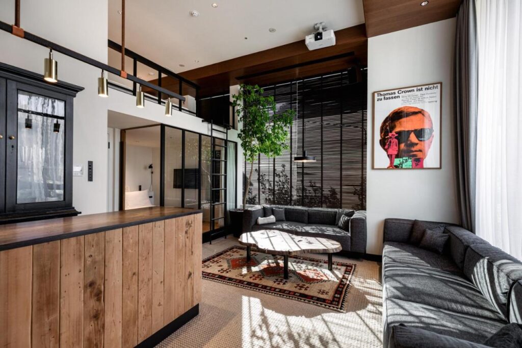 15 Unique Hotels in Tokyo: Coolest hotels 7 Modern living room with large windows, a U-shaped gray sofa, round wooden coffee table, indoor tree, and a colorful portrait—reminiscent of the chic style found in unique hotels in Tokyo. Wood accents and high ceilings enhance the natural light.