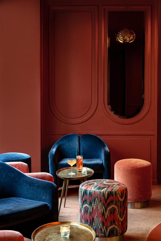 13 Unique Hotels in Paris for a Stylish Stay 55 A stylish lounge in one of the most unique hotels in Paris, with deep red walls, blue velvet chairs, a round gold side table with drinks, and patterned ottomans; ambient lighting creates a cozy, sophisticated atmosphere.