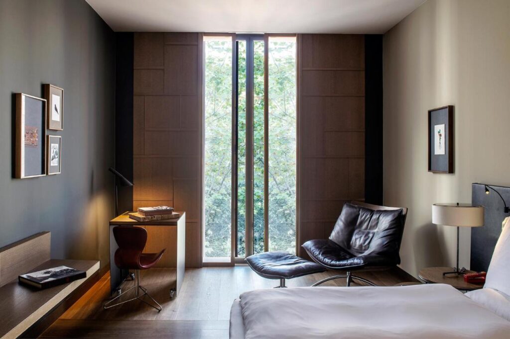 13 Unique Hotels in Barcelona, Spain 3 A modern hotel room in one of the unique hotels in Barcelona, featuring a large bed, leather lounge chair and ottoman, wooden desk with lamp and chair, framed art on the walls, and tall narrow windows overlooking green trees outside.