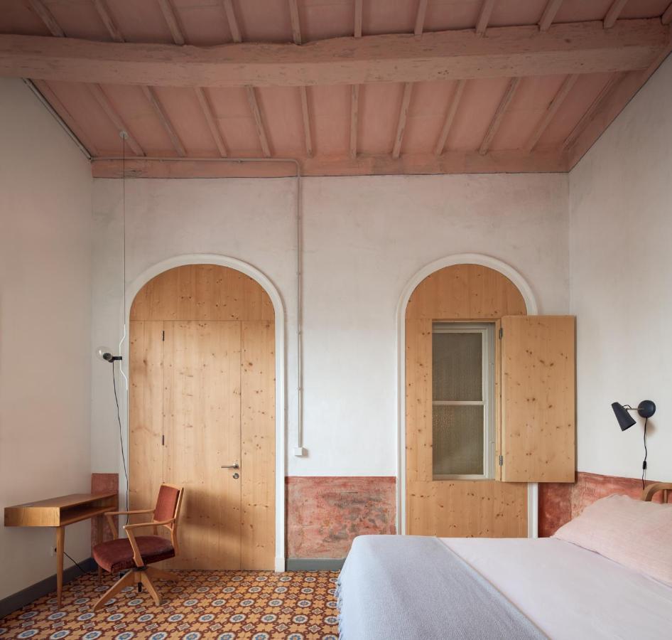 Unique Boutique Hotels in Menorca 15 A cozy bedroom inspired by boutique hotels in Menorca, featuring a patterned tile floor, wooden arched doors, a wooden desk and chair, white and pink bedding, and a high ceiling with exposed beams painted light pink.