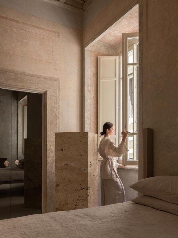 13 UNIQUE Boutique Hotels in Rome 31 A woman in light clothing stands by a tall window in a softly lit, beige-toned bedroom with stone walls—reminiscent of boutique hotels in Rome—looking outside while touching the window frame.