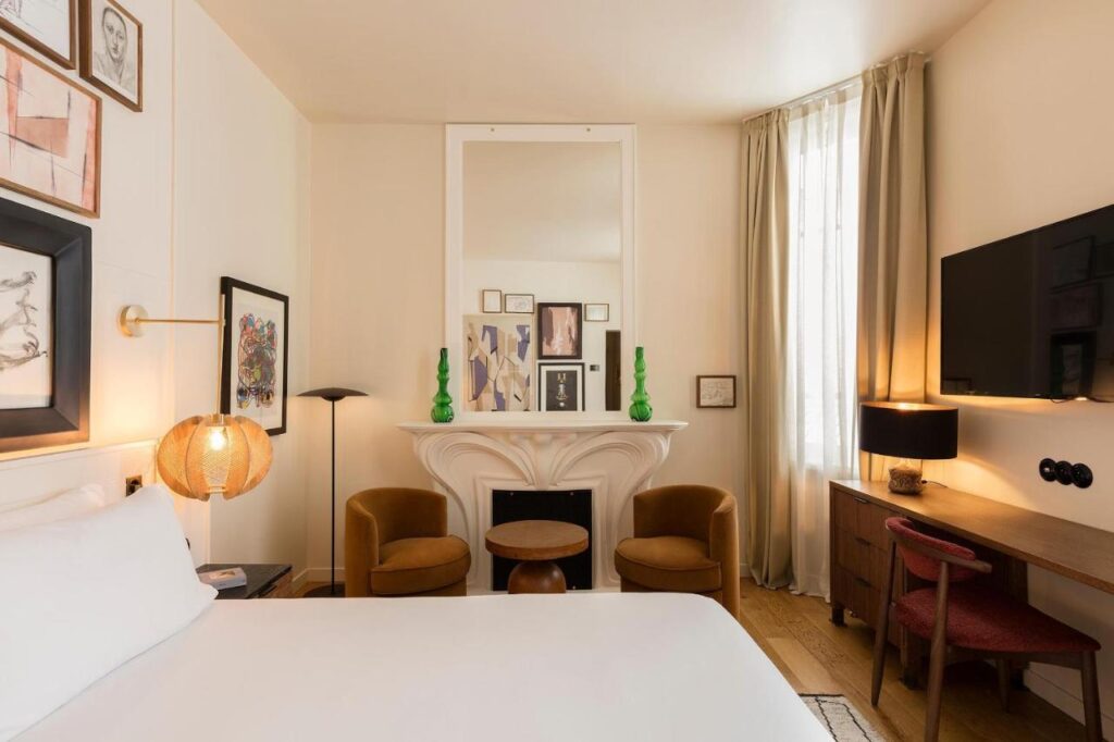 13 Unique Hotels in Paris for a Stylish Stay 23 A cozy, stylish room at one of the unique hotels in Paris, featuring a bed in the foreground, two brown armchairs by a white mantel, a large wall mirror, framed art, a desk with lamp, TV on the wall, and floor-length curtains over a tall window.
