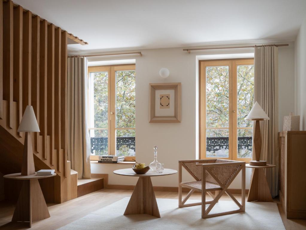 13 Unique Hotels in Paris for a Stylish Stay 28 A modern living room with light wood furniture, a round coffee table, woven chair, and two side tables with lamps evokes the cozy elegance found in unique hotels in Paris. Large windows with beige curtains fill the space with natural light and neutral tones.