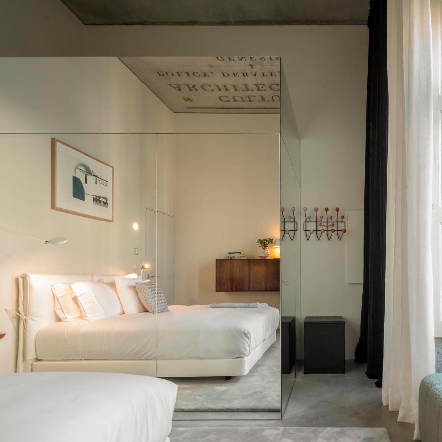 Top 12 Boutique Hotels in Porto, Portugal 17 Minimalist bedroom with a large mirror wall reflecting the bed, white linens, soft lighting, modern artwork, a wooden cabinet, and a wall-mounted coat rack—evoking the serene style of boutique hotels in Porto. Sheer curtains let in natural light from a tall window.
