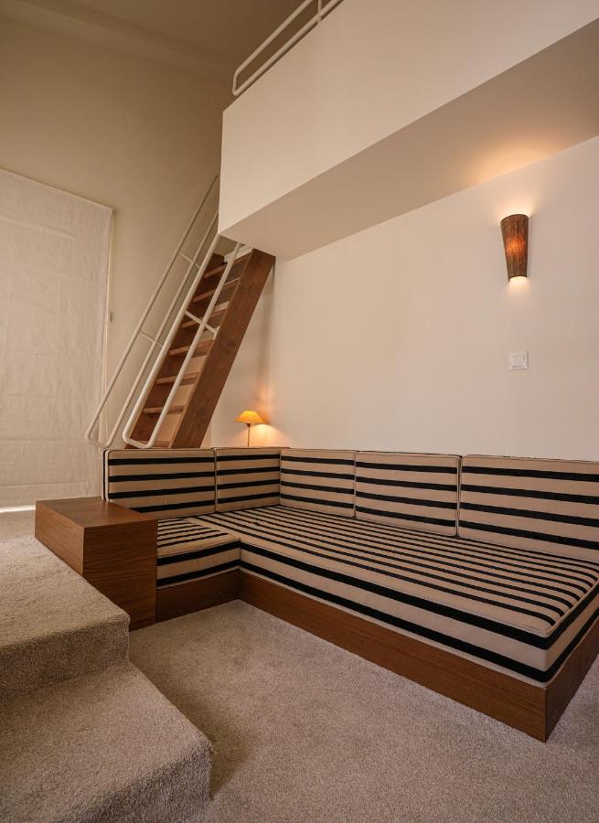A modern loft reminiscent of the best boutique hotels in Athens, featuring a striped L-shaped sofa, beige carpeted floor, wooden stairs to an upper level, a wall-mounted lamp, and minimal decor in a well-lit space.