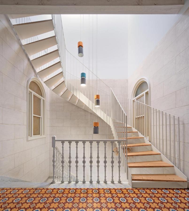 Unique Boutique Hotels in Menorca 14 A modern staircase with wooden steps and sleek metal railings curves upward in a bright, spacious interior—reminiscent of boutique hotels in Menorca—with tiled floors, arched windows, and colorful hanging pendant lights.