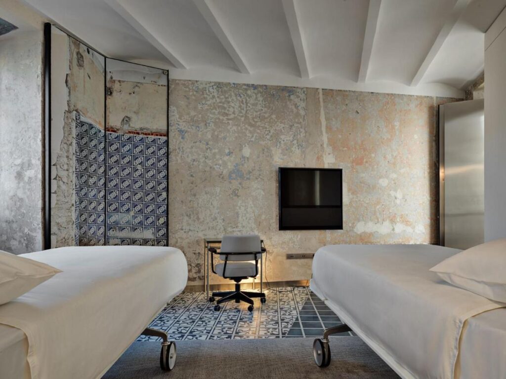 13 UNIQUE Boutique Hotels in Rome 34 Minimalist bedroom with two single beds on wheels, a central desk and chair, patterned floor tiles, a wall-mounted TV, and a textured, partially tiled wall—channeling the chic style of boutique hotels in Rome.