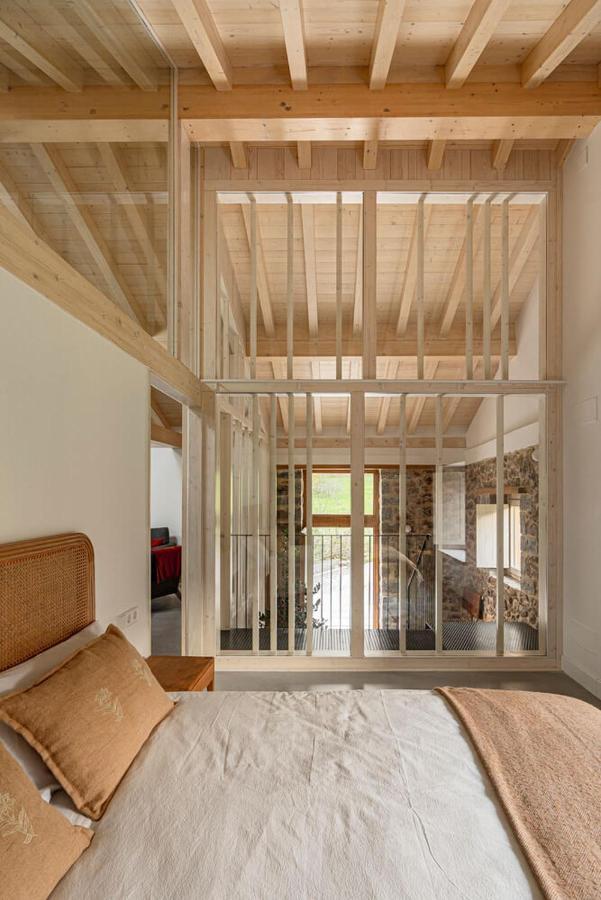 A cozy bedroom in the style of hotels in Cantabria, with a wooden ceiling and beams, beige bedding, and a large glass wall with vertical wooden slats overlooking a bright, rustic living area with stone walls.
