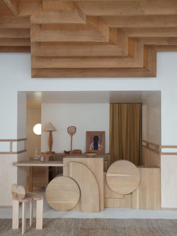 13 Unique Hotels in Paris for a Stylish Stay 27 A modern, minimalist room reminiscent of unique hotels in Paris features light wood furniture with geometric shapes. A countertop, two stools, artwork, and decor sit beneath a wood-paneled ceiling and neutral-toned curtain.