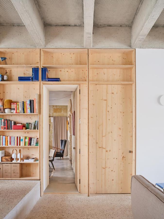 Unique Boutique Hotels in Menorca 36 A modern interior reminiscent of boutique hotels in Menorca features light wooden built-in shelves filled with books and decor. A sliding wooden door and open doorway lead to a hallway, with exposed concrete beams visible on the ceiling.