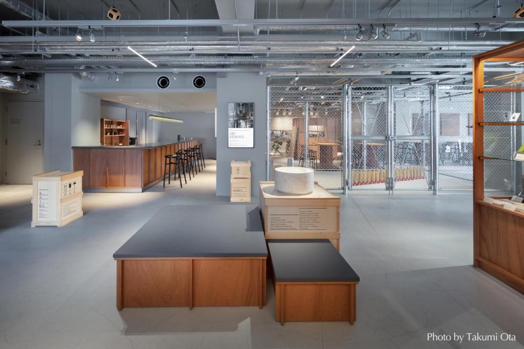 15 Unique Hotels in Tokyo: Coolest hotels 48 Modern, spacious interior with sleek wood and metal furniture, a bar area with stools on the left, display objects in the center, and a wire mesh enclosure on the right—an atmosphere found in unique hotels in Tokyo. Exposed pipes and bright lighting throughout.