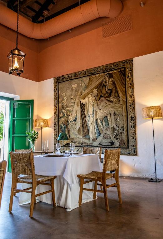 TOP 10 Luxury Hotels in Andalucía 37 A cozy dining area with a white-clothed table set for two, wicker chairs, and a large ornate tapestry evokes the charm of luxury hotels in Andalucía. Warm lighting from lanterns enhances the rustic, artistic ambiance of the room.
