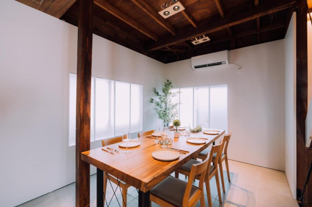 15 Unique Hotels in Tokyo: Coolest hotels 55 A modern dining room with a wooden table set for four, light wood chairs, minimalist decor, large frosted windows, and a potted plant evokes the serene style found in unique hotels in Tokyo. Natural light and an air conditioner brighten the white-walled space.