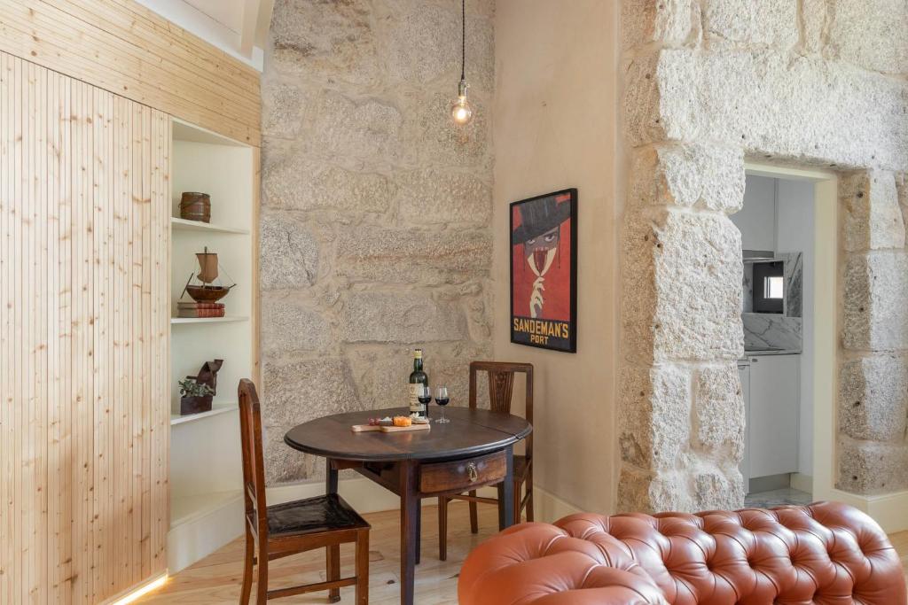 Top 12 Boutique Hotels in Porto, Portugal 43 A cozy room reminiscent of boutique hotels in Porto, with stone walls, a round wooden table set for two with wine and glasses, vintage chairs, a leather sofa, shelves filled with decor, and a poster beside an open doorway to the kitchen.