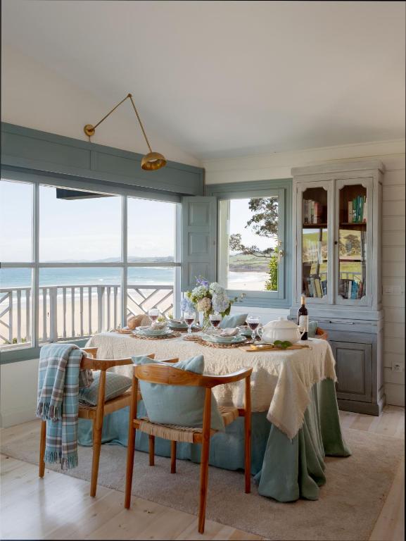 A cozy dining area by large windows overlooking a beach, with a table set for a meal, wooden chairs, and blue and white tablecloth evokes the charm of boutique hotels in Cantabria. Natural light fills the room as flowers decorate the space.