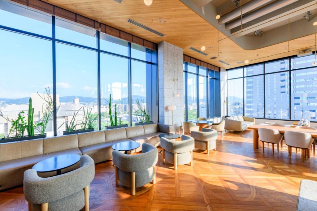 7 Best Hotels in Hiroshima, Japan 13 A modern lounge at one of the best hotels in Hiroshima, Japan, with large windows, light wood floors, round tables, cushioned chairs, and city views. Sunlight fills the spacious room with potted plants along the window ledges.