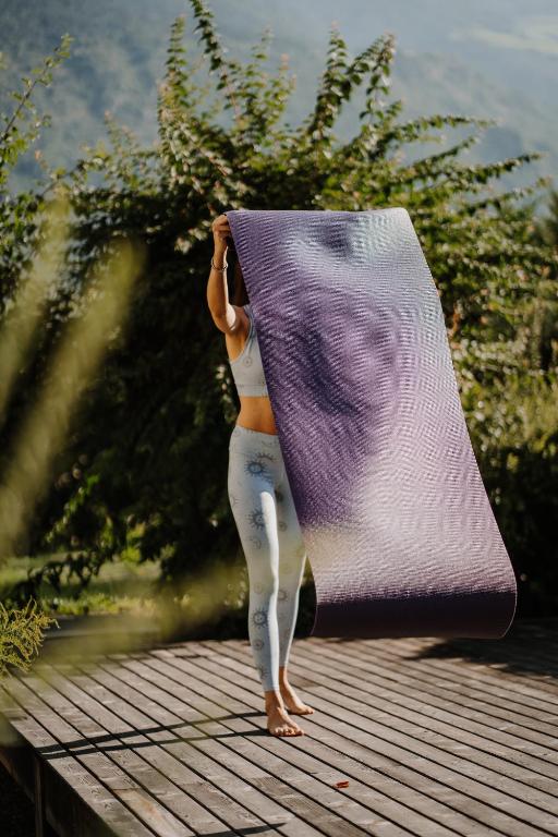 A person in light-colored activewear stands on a wooden deck outdoors, holding up a rolled-out purple yoga mat with green trees and the best hotels in the Dolomites visible among the mountains in the background. Their face is obscured by the mat.