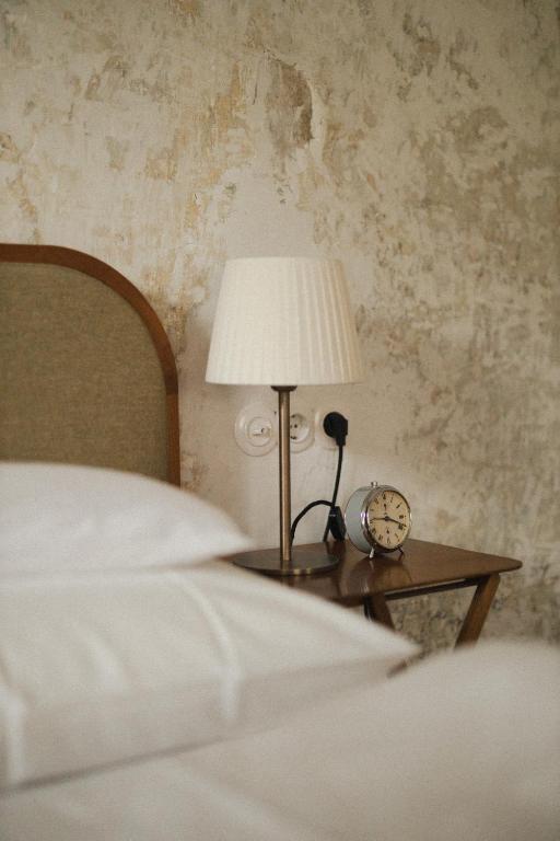 A bedside table with a white lampshade, a classic alarm clock, and an electric socket beside a bed with white sheets against a textured, rustic wall—reflecting the cozy charm found in the best hotels in the Dolomites.