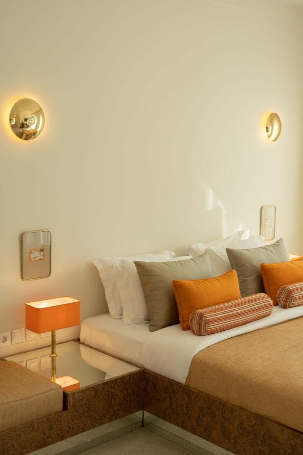 A neatly made bed with tan and white bedding, orange accent pillows, and a matching orange lamp on a mirrored bedside table— reminiscent of the chic style found in the best boutique hotels in Athens. Two round wall lights grace the cream-colored wall above.