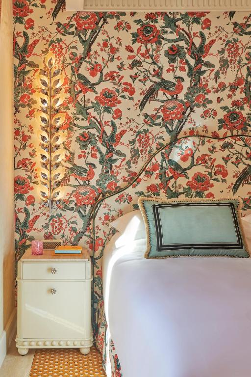 13 UNIQUE Boutique Hotels in Rome 40 A bedside table with books and a glass sits next to a bed with a blue pillow, reminiscent of the charming decor found in boutique hotels in Rome. The floral wallpaper and leaf-shaped wall sconce add warmth and elegance to the space.