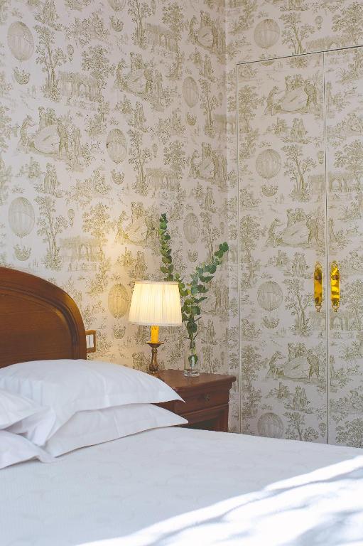13 Unique Hotels in Paris for a Stylish Stay 41 A cozy bedroom corner inspired by unique hotels in Paris, with a wooden headboard, neatly stacked white pillows, a bedside table holding a lamp and a vase with greenery, and light-patterned wallpaper with decorative gold-handled closet doors.