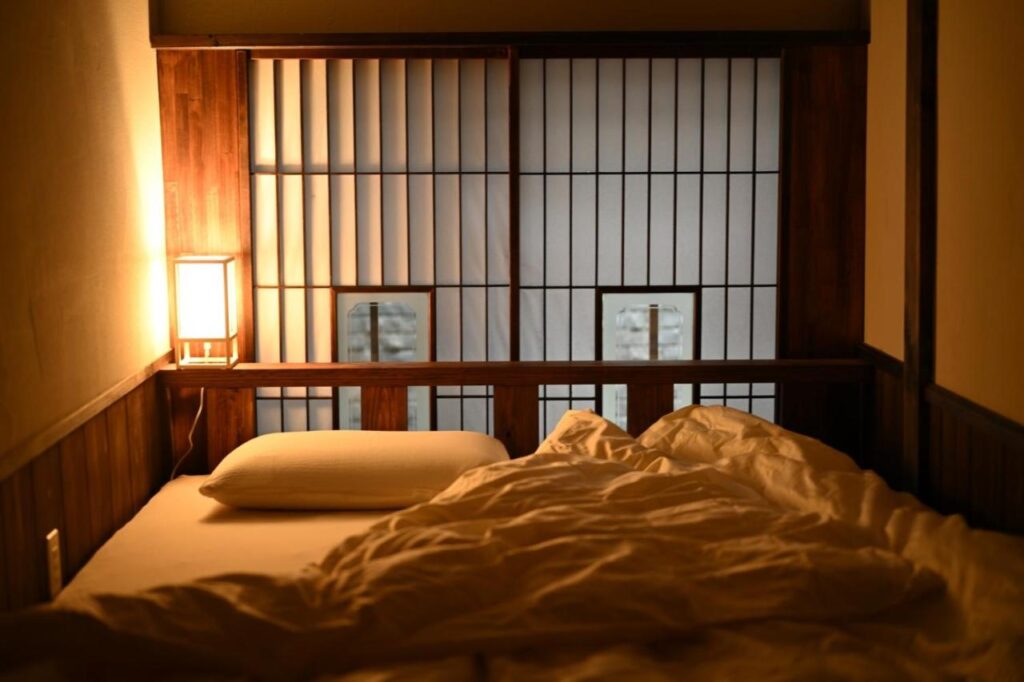 15 Unique Hotels in Tokyo: Coolest hotels 18 A cozy bed with white bedding and a pillow is set next to wooden shoji screens, softly lit by a warm lamp in a small, tranquil room with Japanese-inspired decor—perfect for those seeking unique hotels in Tokyo.