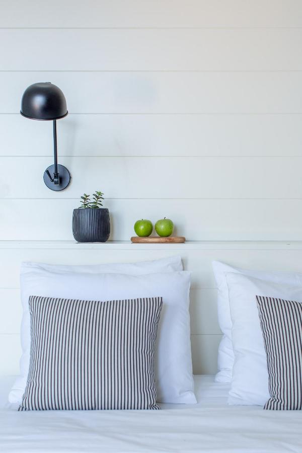Boutique Hotels in Korčula Island 3 A neatly made bed with white pillows and striped cushion covers, reminiscent of boutique hotels in Korcula. Above the bed are a black wall lamp, a small succulent plant in a dark pot, and two green apples on a wooden tray against a white paneled wall.