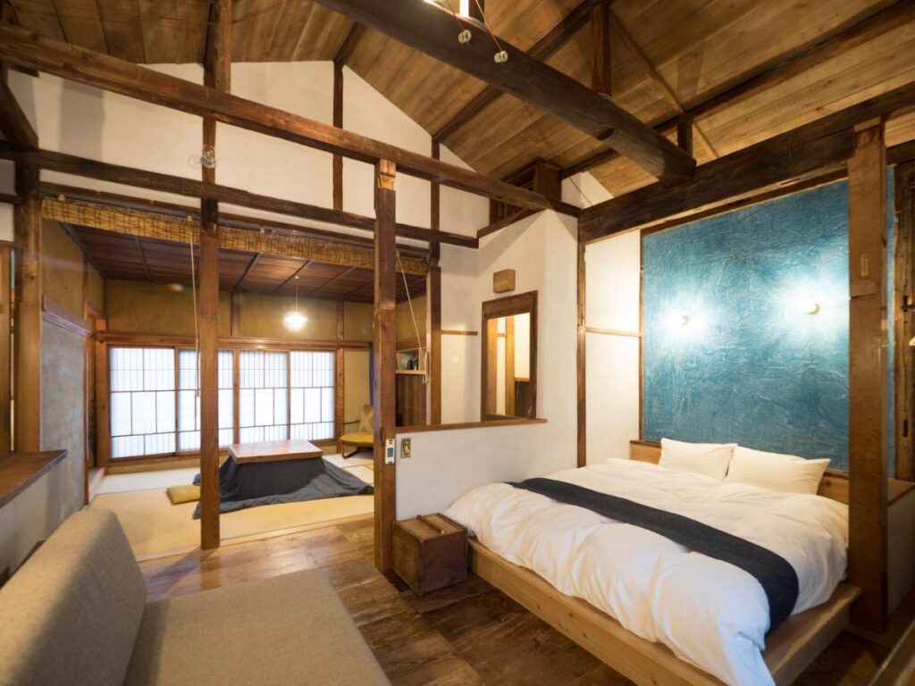 15 Unique Hotels in Tokyo: Coolest hotels 53 A cozy Japanese-style room in one of the unique hotels in Tokyo, with wooden beams, a bed with white bedding, a blue accent wall, and a tatami mat area featuring a low table and floor cushions, softly lit by natural light from shoji screens.