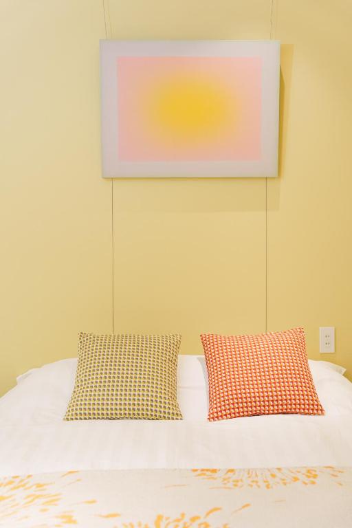 15 Unique Hotels in Tokyo: Coolest hotels 58 A neatly made bed with two patterned pillows, one yellow and one orange, sits against a light yellow wall. Above the bed hangs a soft, square artwork with a pink and yellow gradient—an inviting scene found in unique hotels in Tokyo.