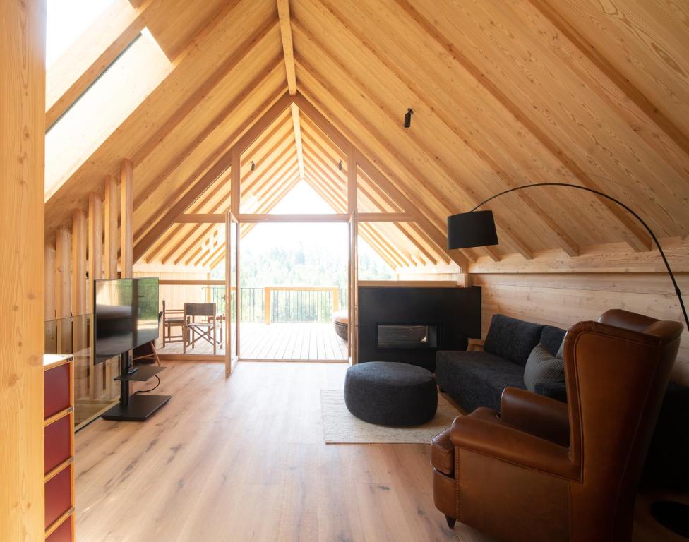 A cozy attic living room with wooden walls and ceiling, like those found in the best hotels in the Dolomites, featuring a brown armchair, black sofa, round ottoman, floor lamp, TV, and large glass doors opening to a sunlit balcony with outdoor seating.