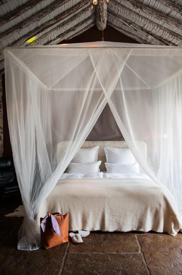 TOP 10 Luxury Hotels in Andalucía 3 A cozy bed with white linens and pillows is covered by a sheer canopy in a rustic room, reminiscent of luxury hotels in Andalucia. A brown bag and white slippers are placed on the floor at the foot of the bed.