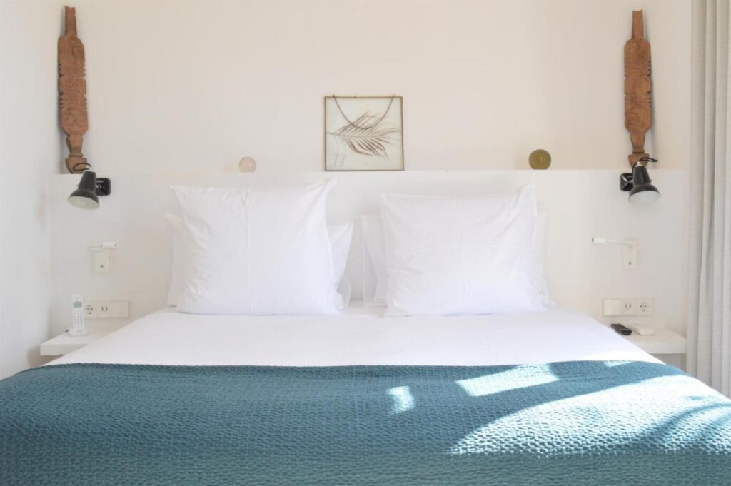 Unique Boutique Hotels in Menorca 28 A neatly made bed with large white pillows and a teal blanket in a bright room. Minimalist decor, like two wall lamps and wooden carvings, creates the calm elegance found in boutique hotels in Menorca, with leaf artwork above the bed.