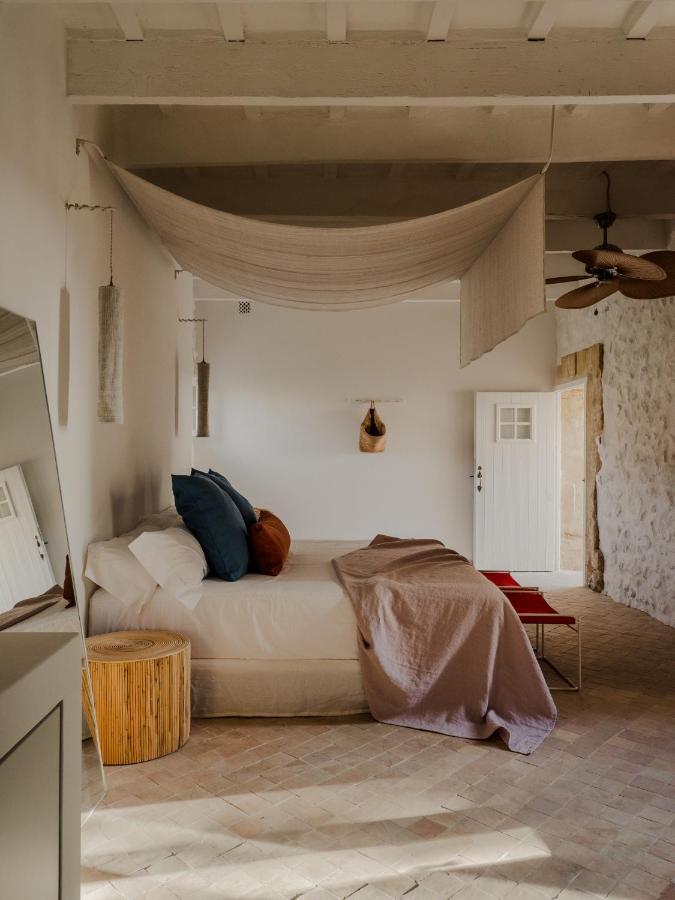 Unique Boutique Hotels in Menorca 18 A cozy bedroom with neutral tones, inspired by boutique hotels in Menorca, featuring a bed with white and earth-tone linens, a hanging fabric canopy, woven side table, stone walls, and a door letting in natural light.
