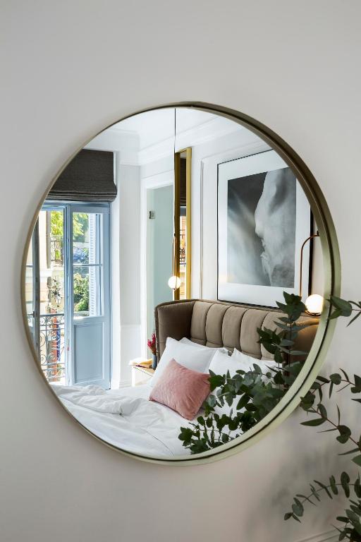 Round mirror reflecting a cozy bedroom reminiscent of boutique hotels in Athens, with a made bed, pink pillow, artwork above the headboard, plants, and an open window letting in natural light.