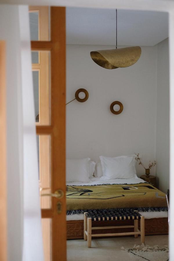 10 Unique Riads in Marrakech with Pool 32 View through an open door into a minimalist bedroom with white walls, a bed with white pillows and a mustard yellow blanket—inviting as some riads in Marrakech with pool—two round wall decorations, and a modern gold light fixture overhead.