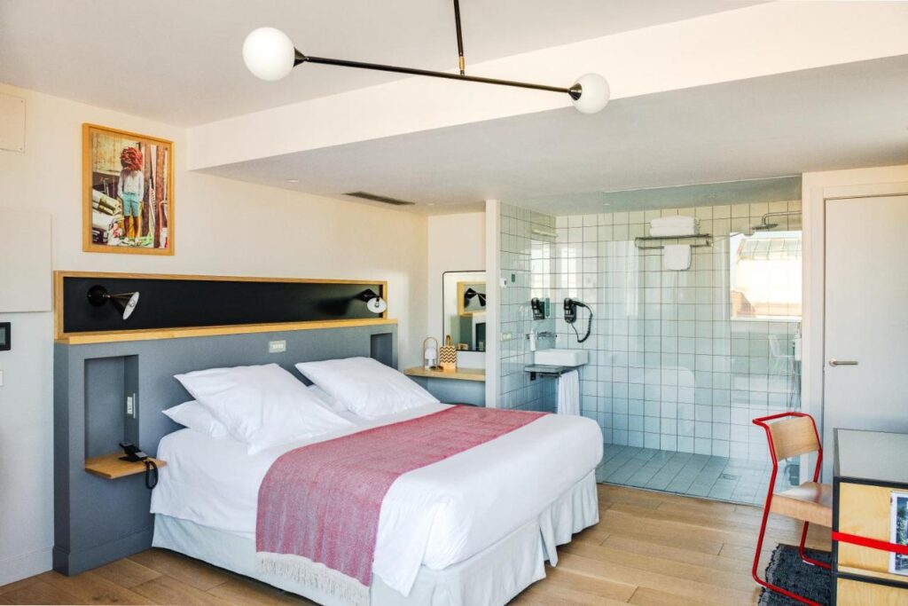 13 Unique Hotels in Barcelona, Spain 38 Bright hotel room with a double bed, crisp white bedding and pink throw, a modern light fixture, a desk with red chair, and an open bathroom area—perfect for those seeking unique hotels in Barcelona.