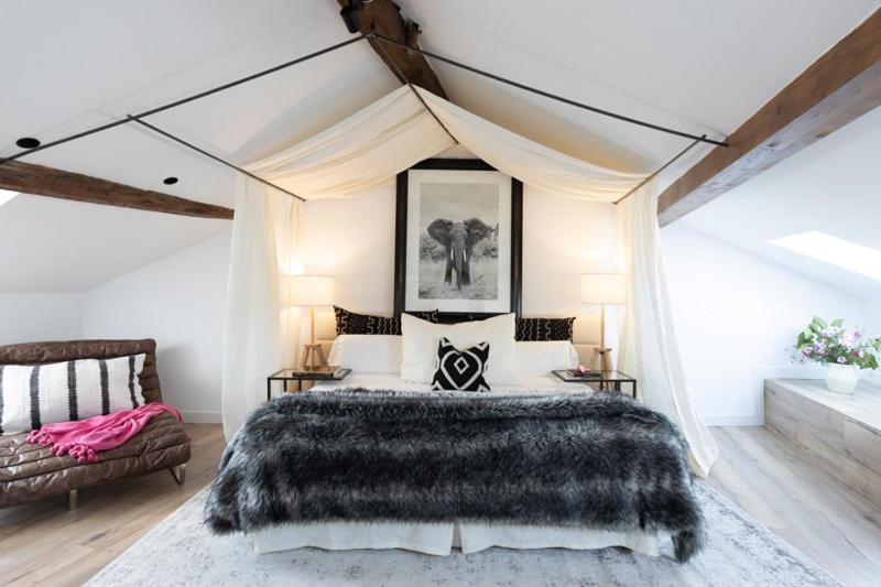 A cozy bedroom reminiscent of boutique hotels in Cantabria, with exposed wooden beams, a canopy bed draped in white fabric, fur blanket, black-and-white pillows, bedside lamps, a framed elephant print, and a brown chair with a pink blanket.