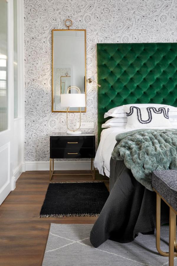 Unique Places to Stay in Cape Town 10 A stylish bedroom reminiscent of unique places to stay in Cape Town, featuring a green tufted headboard, patterned wallpaper, a black nightstand with gold accents, a tall mirror, white lamp, layered bedding, and a textured gray throw blanket.