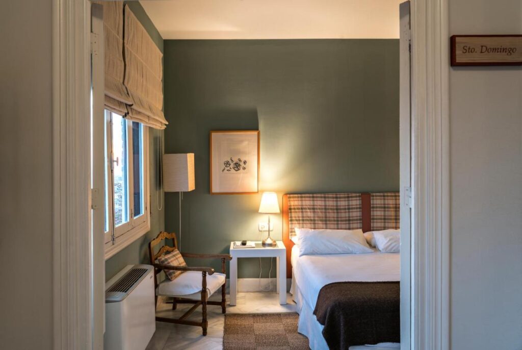 A cozy bedroom inspired by boutique hotels in Granada, featuring a double bed with a plaid headboard, bedside table with a lamp, framed artwork, an armchair, and a window with roman shades letting in natural light.