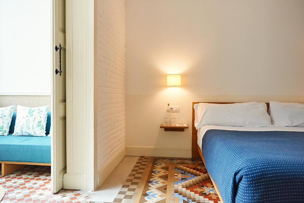 13 Unique Hotels in Barcelona, Spain 51 A modern, bright hotel room at one of the unique hotels in Barcelona, featuring a double bed with a blue blanket, patterned tile floor, wall lamp, small shelf with water bottles, and a cozy seating area with floral pillows.