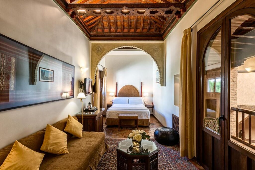 A cozy, elegant bedroom reminiscent of boutique hotels in Granada, with a wooden beamed ceiling, arched entryway, large bed, golden sofa with cushions, coffee table, wall art, and large windows with gold curtains letting in natural light.