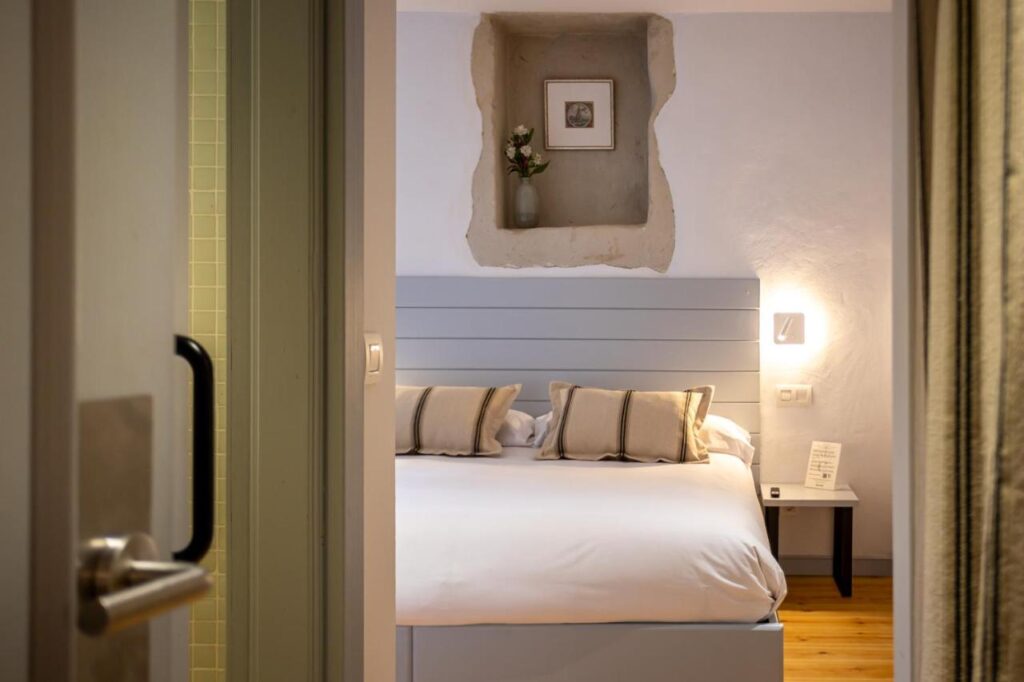 View through an open doorway into a cozy bedroom at one of the charming hotels in Cantabria, featuring a neatly made double bed, four pillows, a small wall lamp, a bedside table with a note, and a decorative alcove above with art and a vase.