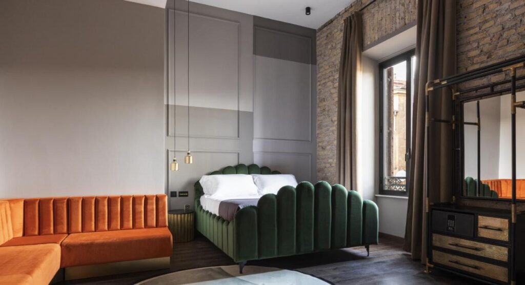 13 UNIQUE Boutique Hotels in Rome 11 A stylish bedroom inspired by boutique hotels in Rome, Italy, with a green scalloped bed, white bedding, orange velvet sectional sofa, exposed brick wall, large window with brown curtains, glass table, and modern gold accents.