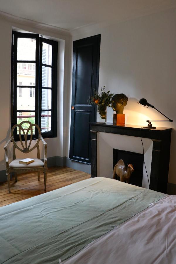 13 Unique Hotels in Paris for a Stylish Stay 14 A bedroom in one of the unique hotels in Paris, featuring a neatly made bed, a fireplace adorned with a lamp, flowers, and sculptures, a wooden chair by black-shuttered windows, and natural light streaming into the room.