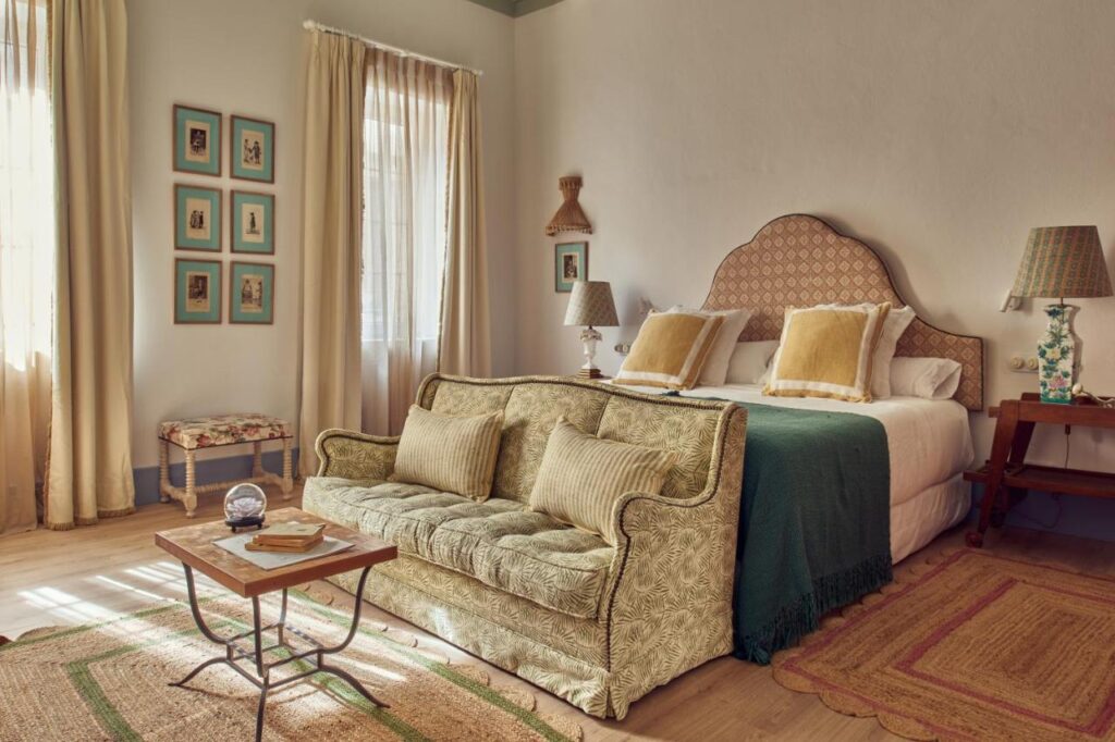 Unique Boutique Hotels in Menorca 7 A cozy, sunlit bedroom inspired by boutique hotels in Menorca features a double bed with patterned headboard, yellow pillows, a green throw, vintage-style sofa at the foot, wooden side tables with lamps, framed art, and beige curtains.