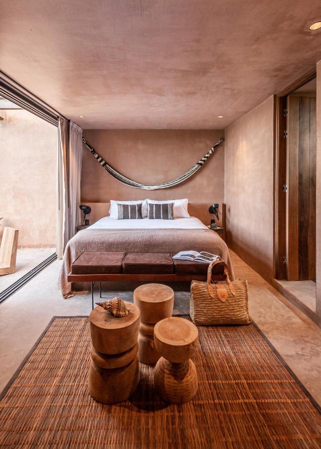 Top Hotels in Tulum, Mexico: Luxury and Beachfront Stays 3 A cozy, modern bedroom with earthy tones—like those in top hotels in Tulum—features a large bed, hammock above the headboard, wooden stools, a woven rug, and wicker basket, with floor-to-ceiling windows letting in natural light.